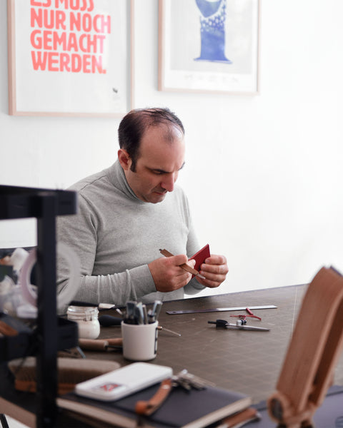 Emre Telli, Gründer und Creative Director von ELBHAVN, an seinem Schreibtisch mit Werkzeugen und Skizzen in einer hellen Atelierumgebung handwerklich tätig ist und sich auf Lederarbeiten konzentriert.