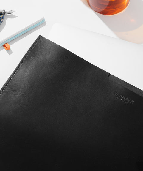 Schwarze MacBook Hülle "Vacchetta Sleeve" für MacBooks auf einem Tisch mit Schreibutensilien und Getränk. Draufsicht.